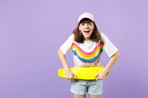 Verrast tienermeisje in levendige kleding met geel skateboard, mond open houden geïsoleerd op violet pastel muur achtergrond in studio. Mensen oprechte emoties, lifestyle concept. Bespotten kopie ruimte.