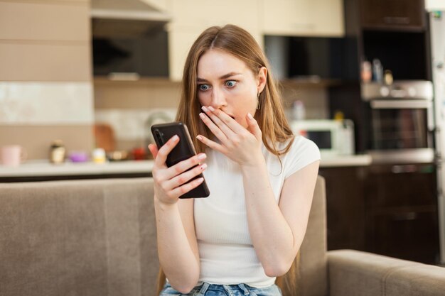 Verraste emotionele vrouw die naar het smartphonescherm kijkt terwijl ze op de bank in de woonkamer zit