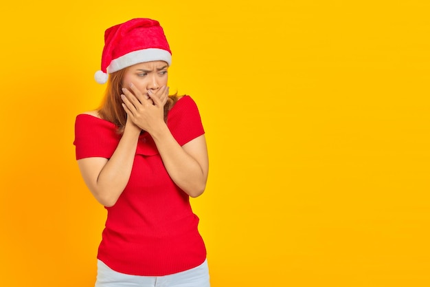 Verraste jonge Aziatische vrouw die een kerstmuts draagt die de mond bedekt met handen op een gele achtergrond