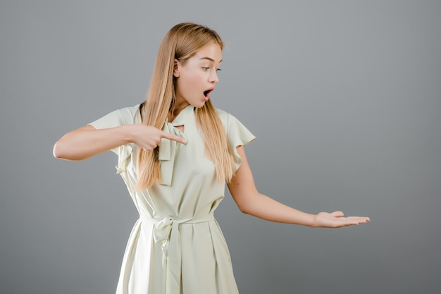 Verraste jonge blondevrouw die vinger richten op copyspace op haar hand die over grijs wordt geïsoleerd