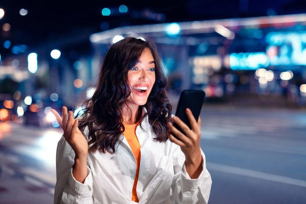 Verraste jonge vrouw die met opwinding naar het smartphonescherm kijkt terwijl ze door de nachtelijke stadsstraat loopt