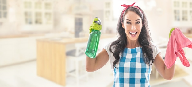 Verraste jonge vrouw in de keuken gekleed in huishoudkleren met een groen wasmiddel en een roze schoonmaakblad.