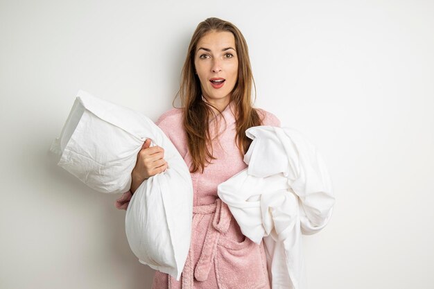 Verraste jonge vrouw in een badjas met een kussen en een laken op een witte achtergrond