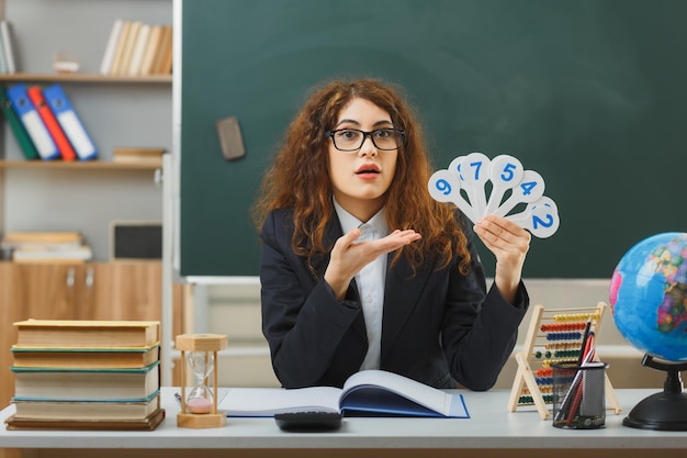 verraste jonge vrouwelijke leraar met een bril die vasthoudt en wijst naar de nummerventilator die aan het bureau zit met schoolhulpmiddelen in de klas