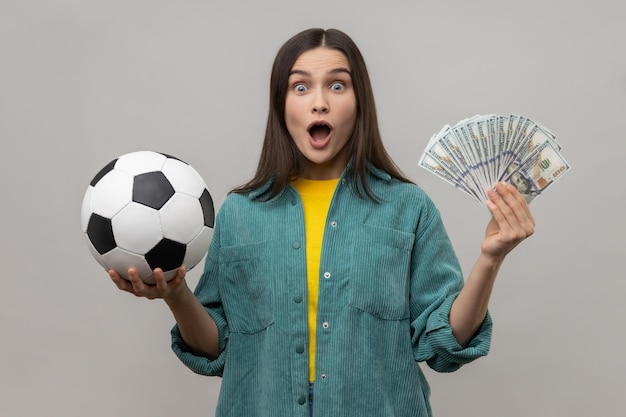 Verraste vrouw die voetbal en honderd-dollarbiljetten laat zien die veel geld wedden voor sport