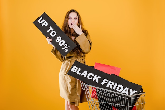 Verraste vrouw in jas met zwarte vrijdag 90% teken en kleurrijke boodschappentassen in winkelwagen geïsoleerd over geel