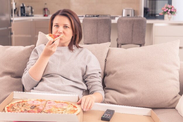 Verraste vrouw pizza eten en tv kijken met afstandsbediening thuis, warme toon