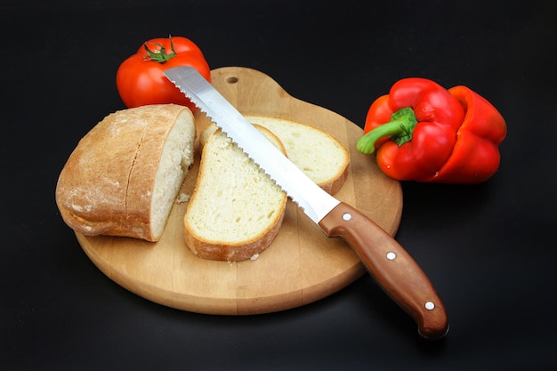 Vers gebakken brood gesneden met een gekarteld broodmes op een houten snijplank met tomaat en peper op zwarte achtergrond