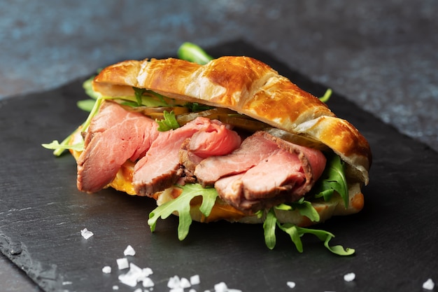 Foto vers gebakken croissantsandwich met rucola en rosbief op een zwarte leisteenplank