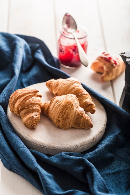 Foto vers gebakken franse croissants met een potje jam op een licht houten tafel met een blauwe servet.