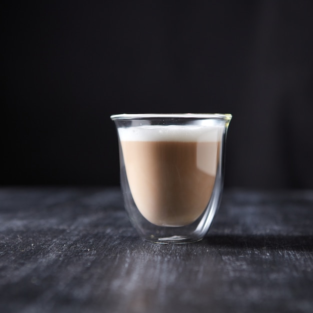 Foto vers gezette cappuccino met schuim in een glazen beker op een zwarte houten tafel rond een donkere achtergrond met kopieerruimte.