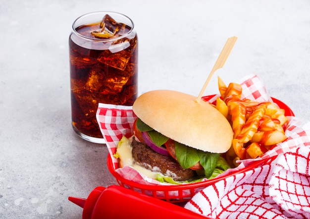 Vers rundvlees hamburger met saus en groenten en glas cola frisdrank met chips frietjes in rode portie mand op stenen keuken.