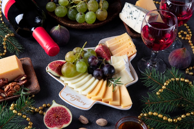 Verscheidenheid aan kaas en fruit geserveerd in plaat als kerstboom, op donkergrijze achtergrond met twee glazen wijn. Snack op oudejaarsavond