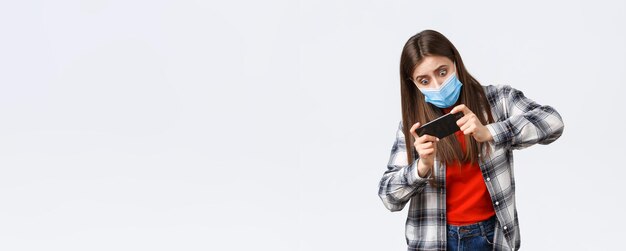 Verschillende emoties covid19 pandemie coronavirus zelfquarantaine en sociaal afstandsconcept Gerichte en vermaakte vrouw met medisch masker die mobiel spel speelt kantelend lichaam moeilijk niveau