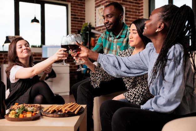 Verschillende etniciteiten vriendengroep, roosteren, wijn drinken, snacks eten, plezier maken op een jong volwassen verjaardagsfeestje in de woonkamer. Multiculturele mensen genieten van vriendschap.