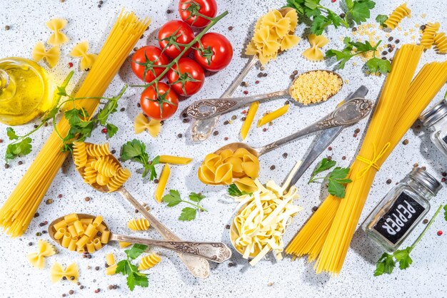 Verschillende gedroogde spaghetti noedels pasta set in lepels op witte tafel achtergrond Verschillende vormen typen rauwe italiaanse pasta met kruiden olijfolie tomaten specerijen voor het koken bovenaanzicht kopie ruimte