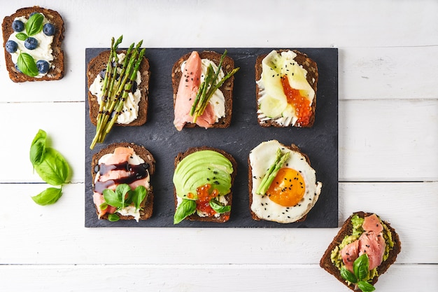 verschillende gezonde sandwiches met verschillende soorten beleg. Vis, kaviaar, avocado, asperges, komkommer, kruiden, sesamzaadjes, glutenvrij brood geserveerd op een donkere schaal