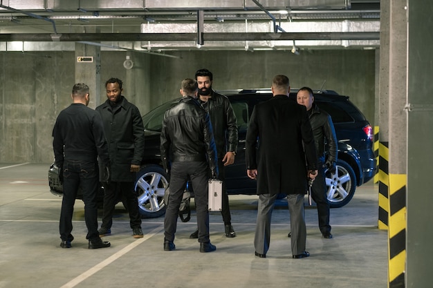 Verschillende interculturele mannen in het zwart houden pistolen en koffer vast terwijl ze de voorwaarden van criminele zaken op parkeerplaats bespreken