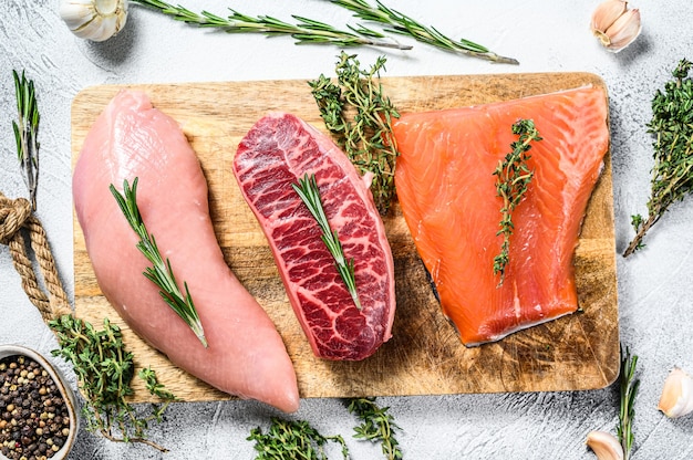 Verschillende soorten rauw vlees op snijplank met kruiden. Runderblad, zalmfilet en kalkoenfilet. steaks. Witte achtergrond. Bovenaanzicht.