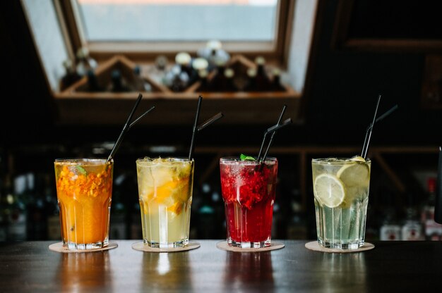 Verschillende soorten zomerdranken in glazen, ijsblokjes en plakjes fruit op donkere wiooden tafel.