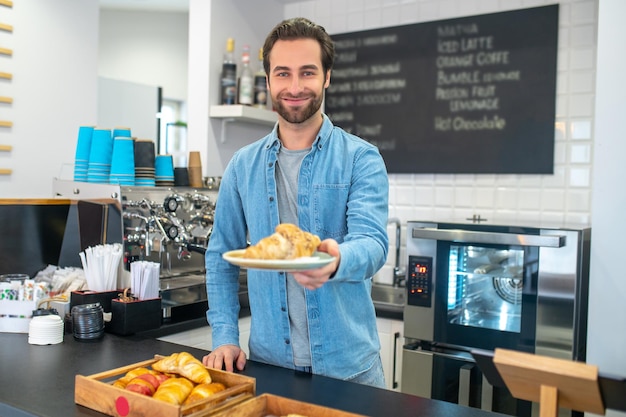 Verse bakkerij. Glimlachende jonge volwassen blanke man die een croissant op een bord steekt naar de camera die aan de toog in café staat