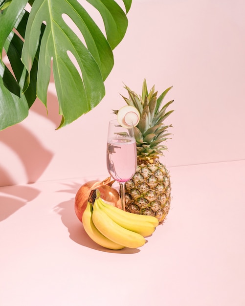 Verse bananen, ananas, granaatappel en verfrissingssap in glas op pastelroze achtergrond.