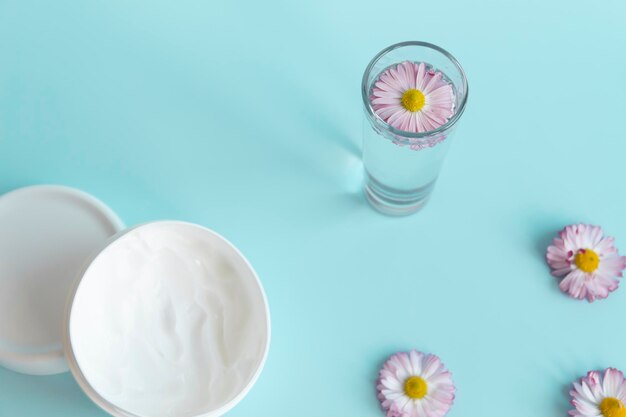 Verse bloemen madeliefjes zijn neergelegd in een hoek met een glas schoon water en een witte pot met een crème cosmetica op een blauwe achtergrond