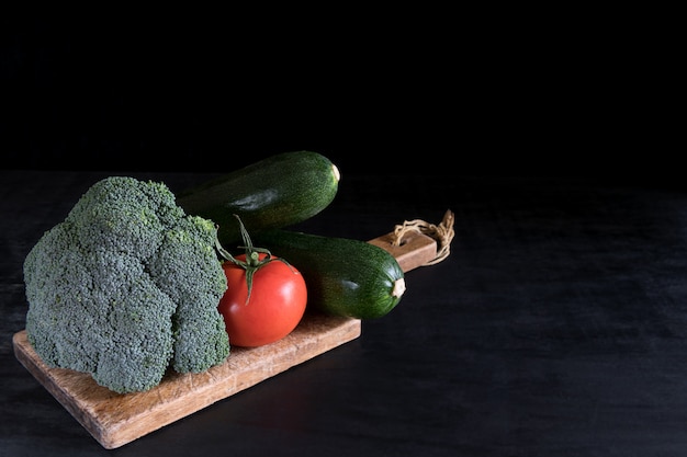 Verse broccoli, courgette en een tomaat op een snijplank op een zwarte tafel, rustieke stijl, donkere sleutel