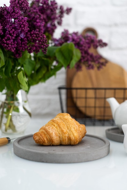 Verse croissant op een grijze plaat op een tafel met een witte theepot en seringen
