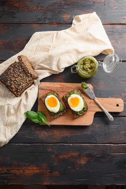 Foto verse groene basilicum pesto in glazen pot zelfgemaakte op oude rustieke houten eieren panini brood met pesto mes en hete sriracha saus italiaans ontbijt met ingrediënten groene pesto bovenaanzicht verticaal