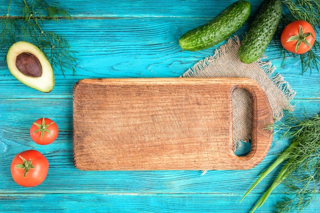 Verse groenten, houten plank en mes op blauwe achtergrond. tomaten, komkommers, avocado, dille en groene ui. ruimte voor tekst.