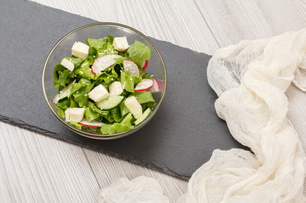 Verse groentesalade met kaas, komkommer en radijs in glazen kom, witte doek op stenen bord. bovenaanzicht.