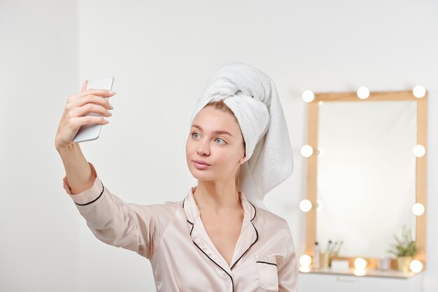 Verse jonge vrouw in zijden pyjama selfie maken na het douchen terwijl je met spiegel en lampen staat