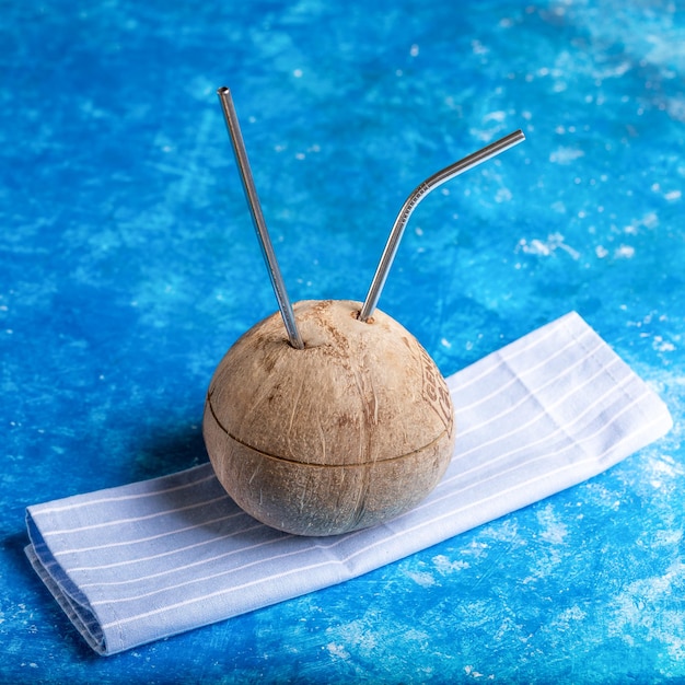 Verse kokosnoot met twee metalen rietjes erin geplaatst op gestreepte keukenhanddoek op blauwe achtergrond