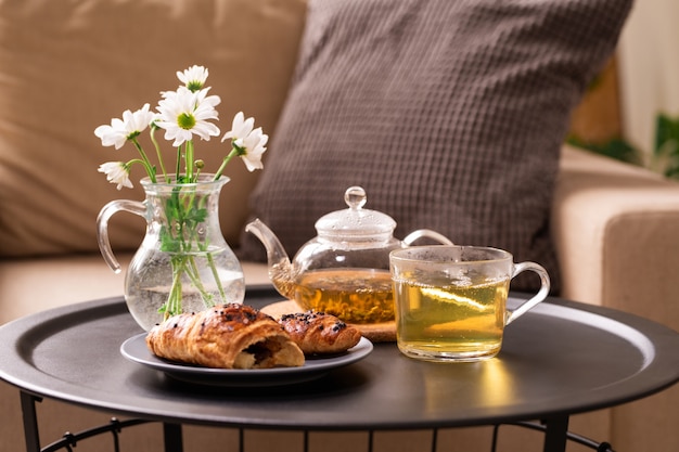 Verse kruidenthee in kop en theepot, zelfgemaakte croissant op plaat en glazen kan met witte bloemen op tafeltje tegen kussen op fauteuil