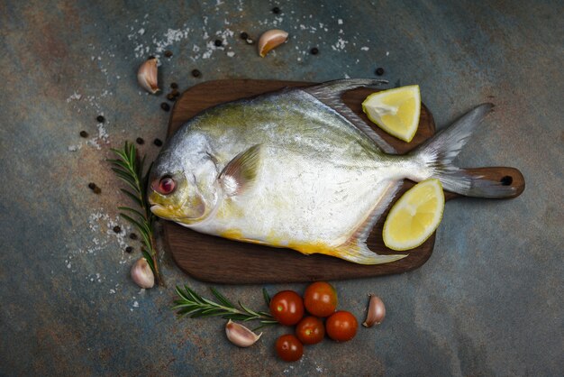 Verse pomfret vis met kruiden specerijen rozemarijn tomaat en citroen op houten snijplank en zwarte plaat achtergrond - Rauwe zwarte pomfret vis