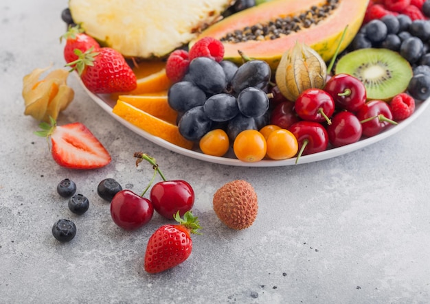 Verse rauwe biologische zomerbessen en exotisch fruit in witte plaat op lichte achtergrond. ananas, papaya, druiven, nectarine, sinaasappel, abrikoos, kiwi, peer, lychees, kers en physalis. bovenaanzicht