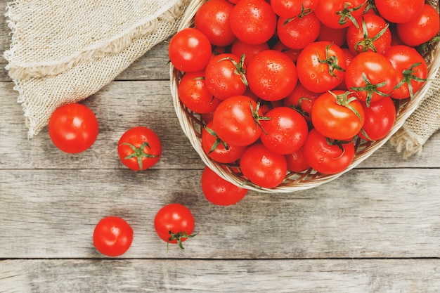 Verse rode tomaten in een rieten mand op een oude houten tafel. Rijpe en sappige kerstomaten met druppels vocht, grijze houten tafel, rond een doek van jute. In een rustieke stijl.