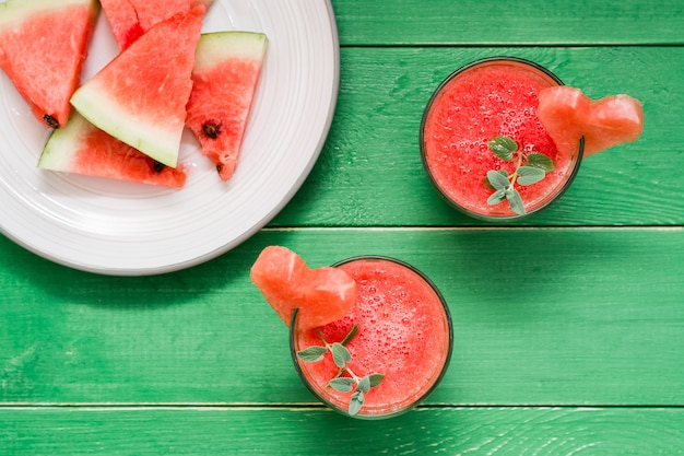 Verse watermeloen gemengd drankje met muntblaadjes en een hart van watermeloen in glazen en plakjes watermeloen op plaat op een houten tafel. Bovenaanzicht