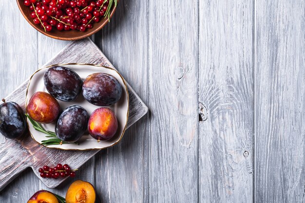 Verse zoete pruimen fruit geheel en gesneden in plaat met rozemarijnblaadjes op oude snijplank met rode bessen bessen in houten kom, grijze houten achtergrond, bovenaanzicht kopie ruimte