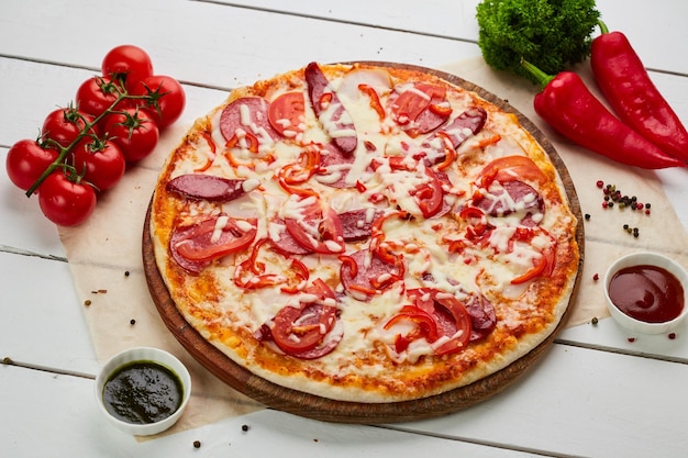 Versgebakken pizza met gerookte worst, rode peper en tomaten geserveerd op houten ondergrond met saus en kruiden voedselleveringsconcept restaurantmenu