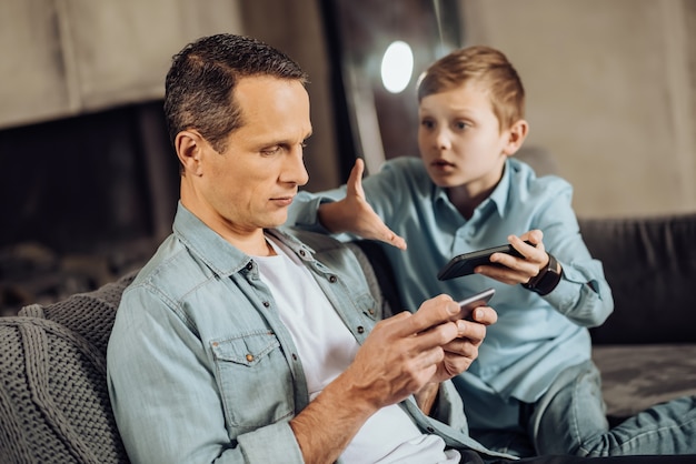 Verslavend gamen. Boos pre-tienerjongen zittend op de bank en probeert de aandacht van zijn vader te trekken door tegen hem te klagen, terwijl de vader verslaafd is aan gamen