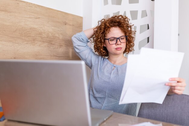 Verstoorde bedrijfsvrouw die documenten en administratie bekijken zit zij bij de lijst bij werkstation. jonge vrouw in haar kantoor die op laptop werkt