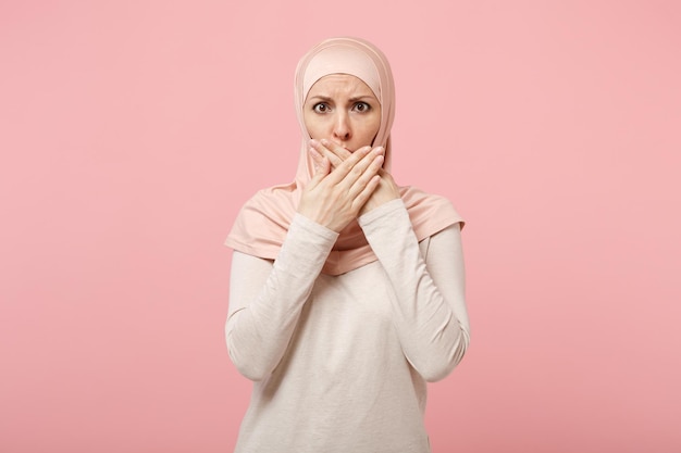 Verstoorde jonge arabische moslimvrouw in hijab lichte kleding poseren geïsoleerd op roze achtergrond studio portret. mensen religieuze islam levensstijl concept. bespotten kopie ruimte. mond bedekken met handen.