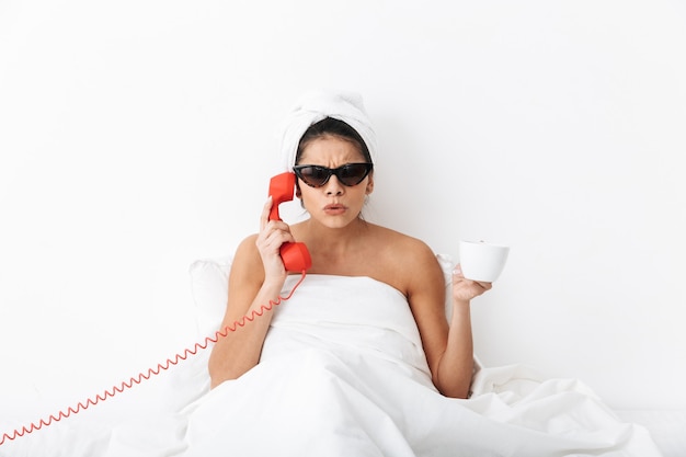 Verstoorde jonge vrouw die na het douchen in bed zit, in een deken gewikkeld, zonnebril draagt, op een vaste telefoon praat, een kopje vasthoudt