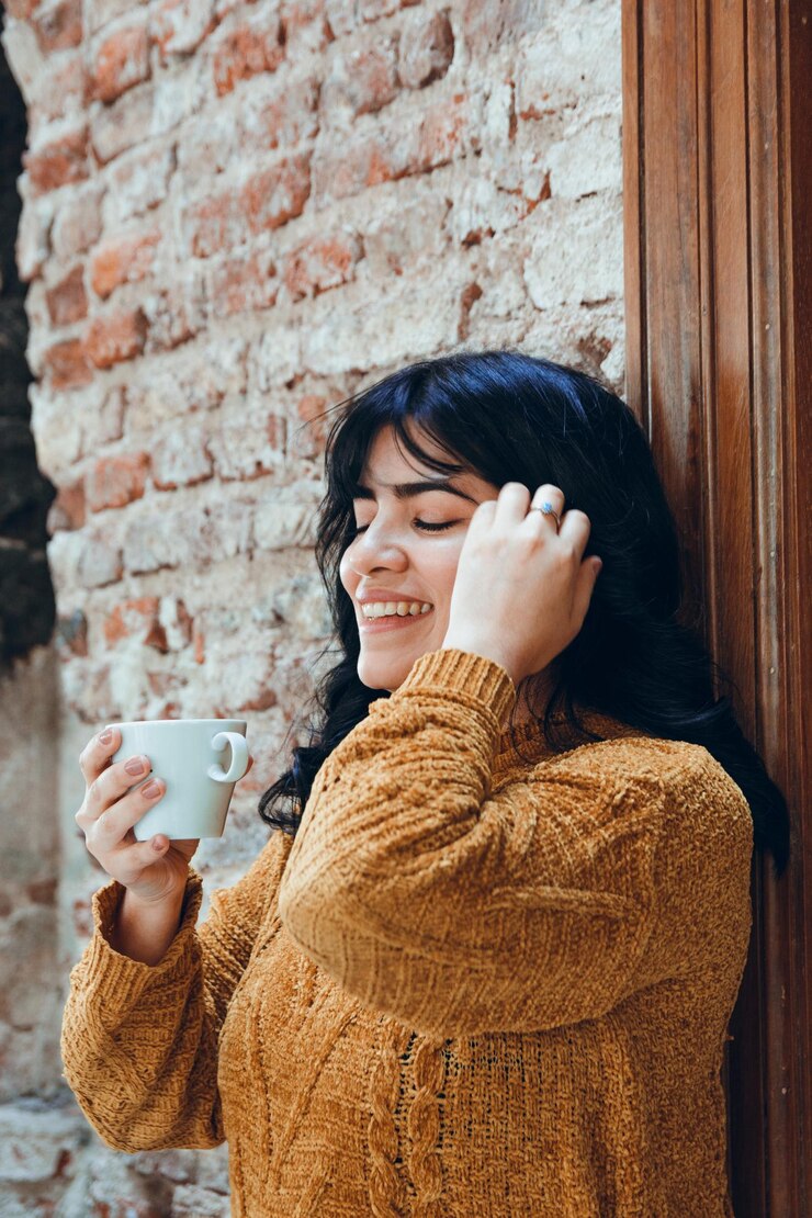 Verticaal beeld van een jonge Latijnse vrouw die alleen tegen de muur leunt met een kop koffie in haar hand.
