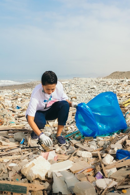 Verticaal beeld van een latijnse vrouw die vrijwillig afval ophaalt op een vervuild strand.