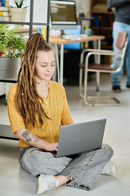 Verticaal beeld van jonge vrouw met dreadlocks zittend op de vloer in kantoor en typend op laptop