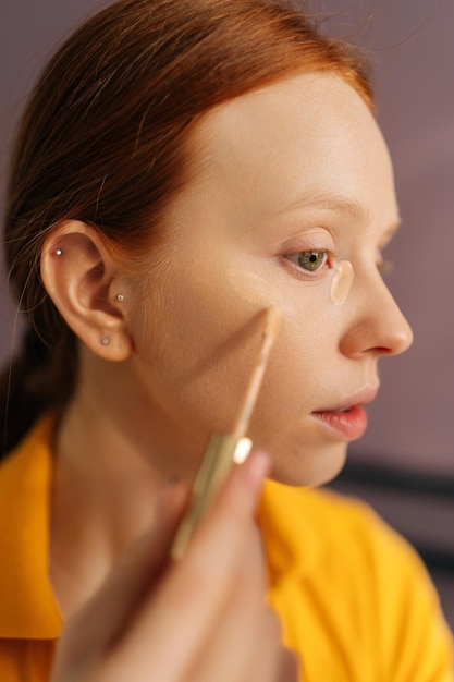 Verticaal close-upportret van mooie jonge vrouw die concealer op het gezicht onder oogcirkels aanbrengt om de huid in de slaapkamer te corrigeren Leuke roodharige vrouw die make-up doet