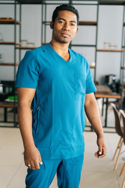 Verticaal medium geschoten portret van een serieuze Afro-Amerikaanse mannelijke arts die een blauw chirurgisch medisch uniform draagt en in het ziekenhuis staat en naar de camera kijkt